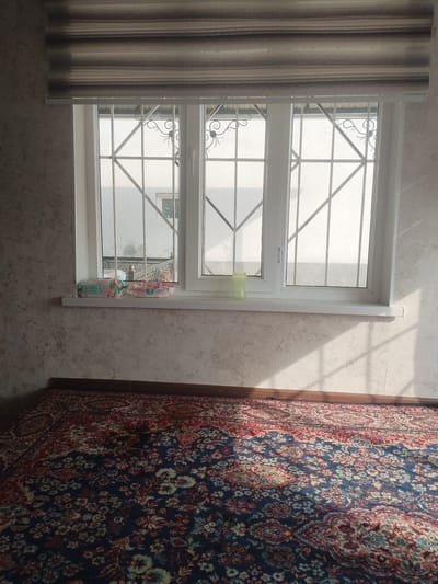 The image shows an interior scene in the city of Октябрь, Kyrgyzstan. The perspective is from a low angle, looking towards a window. The foreground is dominated by a large, ornately patterned rug with a dark blue base and intricate floral designs in red, cream, and other colors. The rug covers the floor and extends upwards towards the wall.
The middle ground features a windowsill against a textured wall. On the windowsill, there are two packages of wet wipes, one with green packaging labeled "Soffia" and another with pink packaging. A single, pale green plastic cup is also visible on the windowsill.
The background consists of a large window with white frames. The window is divided into three panes by vertical and horizontal white mullions. Protective metal bars, decorative in style with curved elements, are mounted on the exterior of the window. Beyond the bars, the outside environment is indistinct and appears bright, suggesting daylight. The light casts strong diagonal shadows from the window bars onto the wall and the rug.
Above the window is a striped roller blind, featuring horizontal bands of varying shades of brown and beige. The overall impression is of a room furnished with a traditional rug, with functional elements like wet wipes and a cup on the windowsill, and security bars on the window.