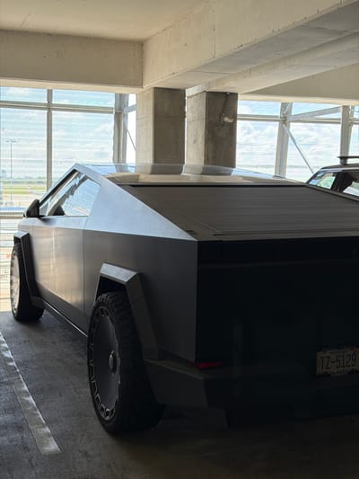 An all-black Tesla Cybertruck is parked indoors, possibly in a parking garage, with a large window in the background revealing a bright, cloudy sky and what appears to be an airport runway or open field. The truck's distinctive angular design is prominent, with its matte black finish and large, unique wheel rims. The rear of the vehicle shows a license plate with the characters "TZ-5129" and text indicating "NORTH CAROLINA CHARLOTTE." Another vehicle, possibly a dark SUV, is partially visible to the right of the Cybertruck. The environment consists of concrete pillars and ceiling beams, suggesting an industrial or urban setting. The lighting is somewhat dim, with light filtering in through the large window. No people are visible in the image.