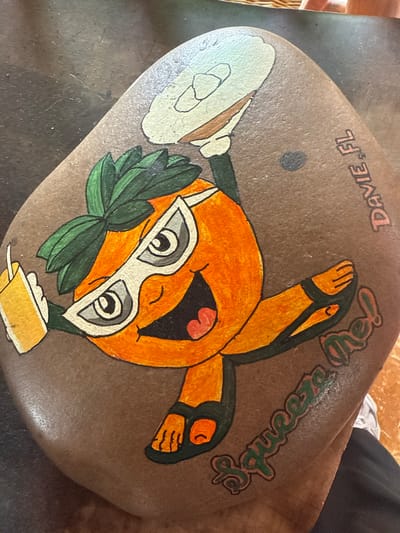 A rock painted with a cartoon orange character wearing sunglasses and holding a drink and a plate is depicted. The words "Squeeze Me!" and "Davie, FL" are also painted on the rock. The character appears to be associated with the city of Davie, Florida, United States of America.