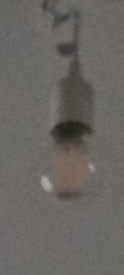 The image shows a dimly lit, out-of-focus view of a light bulb hanging from a wire against a textured, grey surface. The light bulb has a visible filament and a slightly amber-colored glass. There are some small, white specks on the surface, possibly dust or reflections. The background appears to be a wall or ceiling. There is no identifiable information within the image that would place its location in the city of Oldenburg, Germany.