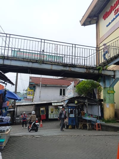 The image portrays an urban street scene in Kidapawan, Philippines, under an overcast sky. A grey-painted steel pedestrian overpass with black railings dominates the upper half of the frame, showing signs of corrosion and minor vegetative growth. A long green banner with white text is visible through the railings on the far side of the overpass. One individual is discernible walking on the far side of the overpass, while another person, dressed in light-colored clothing, is seen ascending the staircase leading to the overpass on the right, adjacent to a yellow two-story building featuring a red sign with partially visible white text. A green and yellow sign reading "WC/MUSHOLA" with a right-pointing arrow is affixed to the overpass's support column on the right.
At street level, a paved surface, resembling cobblestones, serves as a thoroughfare and commercial area. On the left, multiple makeshift vendor stalls are present, one shaded by a blue umbrella and another displaying a "CIPANAS" sign. A person in a red and blue uniform stands near an orange traffic cone. A second individual, wearing a white helmet and light-colored jacket, is seated on a black motorcycle, facing away from the viewer.
In the mid-ground, towards the center and right, several street food stalls are operational. A blue vendor cart prominently displays "BUBUR AYAM" (Chicken Porridge) in white lettering. Wooden tables and red plastic stools are arranged for seating beside this stall. A man wearing a dark jacket and light shirt, carrying a shoulder bag, walks across the paved area from the right, heading towards the left foreground, passing the "BUBUR AYAM" stall. Behind the street-level vendors, a two-story building with a red corrugated roof and white walls is visible.