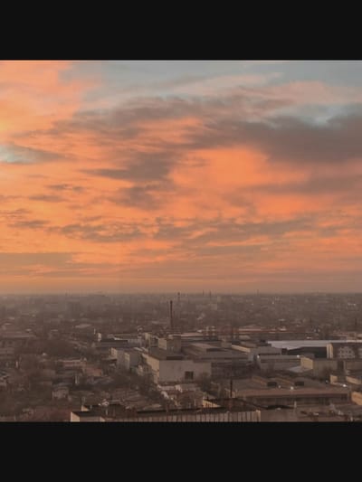 The image depicts a sprawling industrial cityscape at dusk, under a dramatic sky painted with hues of orange, pink, and gray clouds. The foreground features a dense arrangement of industrial buildings, characterized by their flat roofs and uniform structures, interspersed with chimneys. In the distance, the city extends further, with a hazy, low-lying skyline. The overall impression is one of industrial activity and the transition from day to night. There are no discernible people or specific actions taking place in the scene.