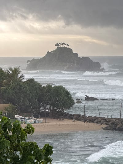 An eye-level, medium shot captures a coastal scene in Ahangama, Sri Lanka, under a hazy, overcast sky. The ocean is active with moderately sized waves crashing against a rocky shoreline. In the middle ground, a small, tree-covered island rises from the sea. Foreground elements include lush green foliage on the left, a sandy beach with two weathered fishing boats, and a breakwater constructed of rocks. The overall atmosphere is moody and somewhat dramatic due to the weather.