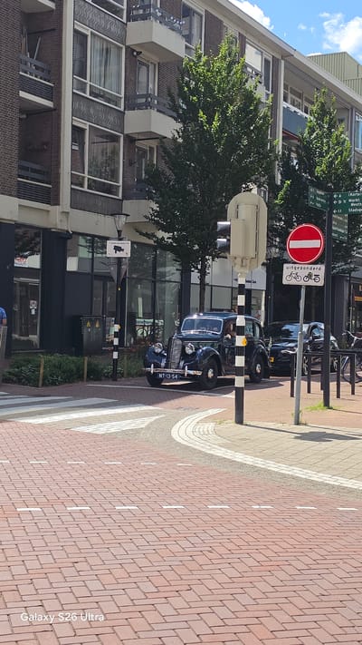 A dark blue vintage car is stopped at a pedestrian crossing on a sunny day in Vlaardingen, Netherlands. The car, a Morris Oxford MO, has a license plate that reads "NT-13-54". The car is facing right, with its headlights on. In the background, there is a modern brick building with balconies and large windows. To the right of the car, there is a traffic light and a street sign indicating directions to various locations, including "Korte Markt" and "Westsingel". A red circular sign with a white horizontal bar is also visible, indicating "no entry". The street is paved with red bricks, and a white pedestrian crossing is marked on the road. The sky is blue with a few scattered clouds.