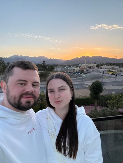An eye-level outdoor selfie captures two individuals, a male and a female, in the foreground at sunset, overlooking the city of Hatta, United Arab Emirates, with a mountainous backdrop. The male, positioned on the left, has dark hair and a beard, wearing a white hooded sweatshirt with "SNRG" in red. The female, on the right, has long dark hair and wears a white hooded sweatshirt with a white shoulder bag strap visible. Both face the camera with neutral expressions. They appear to be on an elevated viewpoint, with a dark railing partially visible in the lower right.
In the midground, the cityscape of Hatta unfolds, featuring a road intersection with several vehicles, including cars and a yellow bus. Low-rise commercial buildings with unidentifiable signage line the roads. A prominent mosque with a single, slender minaret stands out in the center-right background. Sparse green vegetation, including trees and shrubs, is visible throughout the urban landscape.
The far background is dominated by rugged, dark mountains silhouetted against a setting sun. The sun's disk is visible just above the mountains in the center-right, casting a bright orange and yellow glow across the horizon. The sky transitions from vibrant orange and yellow near the mountains to a clear, light blue overhead, with scattered wispy clouds. The scene is illuminated by the soft, warm light of the golden hour.