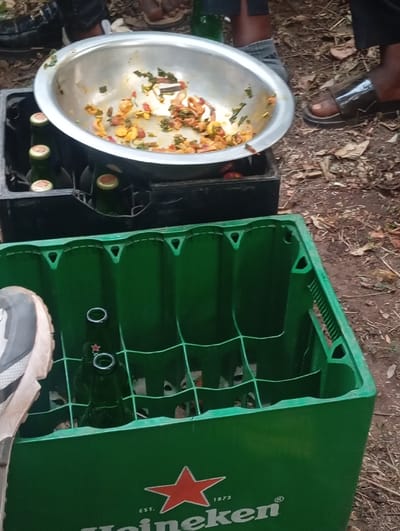 A green Heineken crate holds several empty green beer bottles. A black crate filled with full bottles supports a stainless steel bowl containing a cooked mixture of vegetables and spices. Nearby, several individuals gather, their feet and footwear visible, suggesting a casual outdoor gathering. The scene is set on bare ground covered with sparse vegetation.