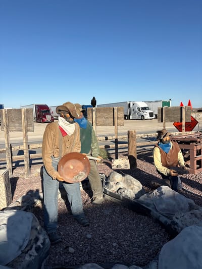 This is a full-shot, eye-level, outdoor photograph that appears to be taken in the daytime under a clear blue sky. The image is oriented vertically. The primary subjects are two mannequins dressed as old-fashioned prospectors, posed in what appears to be a dusty, outdoor exhibit.
The mannequin in the foreground is wearing a brown cowboy hat, a light brown jacket, blue pants, and brown boots. A white and red bandana is tied around its neck. It is holding a round, copper-colored pan, possibly a gold pan, in its hands, with what appears to be rocks or gold nuggets inside. The mannequin behind it is dressed in a yellow shirt, a brown vest, and blue pants, also wearing a blue bandana around its neck. It is seated and appears to be holding a shovel or a similar tool. The mannequins are positioned on a surface covered with small gray rocks and larger, light-colored, irregular-shaped objects that resemble boulders or plaster casts.
Behind the mannequins, there is a wooden fence structure and some industrial elements. A white semi-truck and a red semi-truck are visible in the background, suggesting the location might be near a road or a truck stop. A sign with orange and black lettering is partially visible, with the words "ST RIPS" readable. There are also orange traffic cones in the background. The overall scene evokes a historical reenactment or a roadside attraction related to gold prospecting or the Old West. The lighting suggests it is a sunny day.