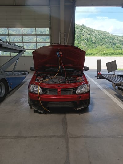 A red car with its hood open is parked on a concrete floor inside a garage or workshop. The car's front bumper is partially removed, exposing its radiator and other components. A blue, red, and yellow manifold gauge set, commonly used for air conditioning system diagnostics or recharging, is connected to the car's engine bay and hanging from the open hood. Hoses from the gauges lead into the engine compartment.
To the left of the red car, part of a large, blue and grey car transport trailer is visible. To the right, another dark-colored flatbed trailer or vehicle structure with a red and white reflective triangular marker is partially seen. The garage has a large, overhead sectional door that is partially open, revealing a vibrant natural landscape outside.
Beyond the garage, a lush, green hillside covered with trees, many of which appear to have white blossoms or light green spring foliage, stretches under a blue sky with some white clouds. The weather outside appears bright and clear, indicating daytime. A metal ladder is leaning against a wall inside the garage on the right side. The concrete floor shows some stains and seams. The text "dci" is faintly visible on a component within the engine bay. The scene depicts a vehicle undergoing service in Dobrodan, Bulgaria.