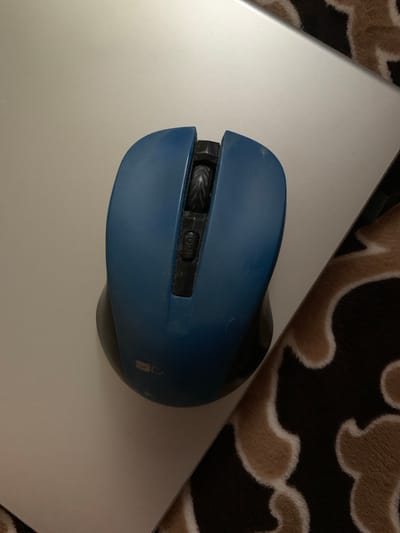 A blue computer mouse is positioned on a light-colored, smooth surface. The mouse has a scroll wheel and two buttons. A textured surface with a brown and cream pattern is visible to the right of the mouse. The setting appears to be indoors, possibly on a desk or table. There is no discernible information about the location in Орлеўскі сельскі Савет, Belarus.