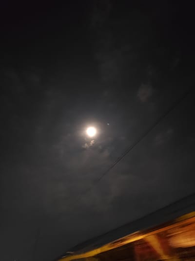 A night view in Lhokmambang, Indonesia, shows a bright full moon partially obscured by thin clouds in a dark sky. A power line diagonally crosses the upper right portion of the frame. At the bottom of the image, there are blurred orange and yellow lights, potentially from a moving vehicle or a building. The overall ambiance is dark due to the night setting.