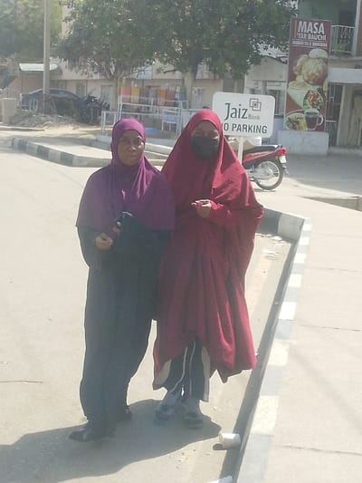 The image captures two adult females standing on a paved sidewalk in Gyadi Gyadi Arewa, Nigeria, under bright daylight. The woman on the left wears a purple hijab and a dark abaya, holding a dark object, and looks towards the camera. The woman on the right wears a deep red khimar, a black face mask, dark pants with a white stripe, and grey/blue shoes, holding a small pink object in her right hand. Behind them, a paved road is visible to the left, separated by a curb painted with black and white stripes. A white "Jaiz Bank NO PARKING" sign and a vertical "MASA YAR BAUCHI" food advertisement banner are present in the mid-ground. A red motorcycle is parked behind the bank sign. Green-leafed trees and a light-colored building are in the background. A dark, possibly damaged vehicle is partially visible in the far left background. A white paper cup lies on the curb near the women's feet.