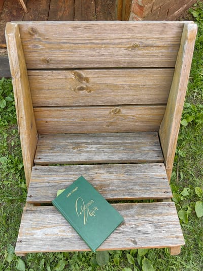 A green book with gold lettering is placed on a rustic wooden chair in an outdoor setting. The chair is made of weathered, light-brown wooden planks, showing signs of age and outdoor exposure. The back of the chair is formed by vertical planks, while the seat is made of horizontal planks. The chair sits on a patch of green grass with some weeds visible. In the background, parts of a wooden structure and a brick wall can be seen. The book, titled "Мартин Иден" (Martin Eden) with the author "Джек Лондон" (Jack London) written above, is open at an angle on the seat of the chair. The lettering on the book is in a decorative script. The scene appears to be a quiet, possibly rural or park-like area. The image does not contain any people or evidence of recent activity. The lighting suggests it is daytime, with soft shadows.