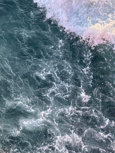 The image displays a high-angle view of turbulent water in the coastal waters near Kato Karioutes, Greece. The water exhibits a deep blue-green hue, with significant areas of white foam and spray indicative of active wave action. Patches of foam in the upper right quadrant show subtle pink and light purple undertones, suggesting reflections from ambient light. The surface is highly agitated with irregular patterns of churning water and numerous whitecaps, consistent with an ocean or sea environment. No individuals, vessels, or man-made objects are visible within the frame. The scene focuses exclusively on the dynamic interaction of water and air.