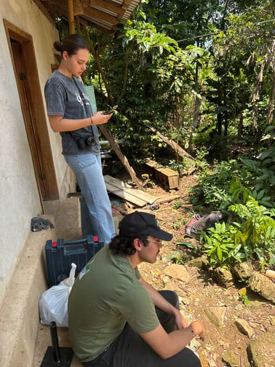 Two individuals are positioned on a concrete structure adjacent to a building with a wooden door and bamboo framing. The female on the left is using a smartphone and has a camera attached to her waist. The male is seated to the right of the female, in front of a hill with vegetation. A black plastic toolbox and a white plastic bag are situated beside the male. The setting is identified as Pinahuista, Mexico.