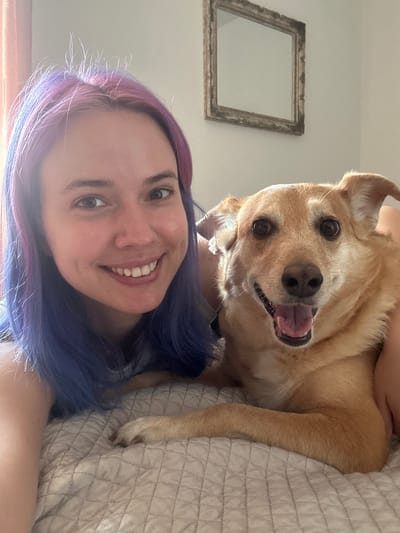 A selfie shows a woman with purple and blue hair smiling at the camera, with a tan dog positioned above her. The dog has its mouth open, tongue visible, and is looking into the camera. They both appear to be lying on a bed with a tan-colored, quilted bedspread. On the wall, slightly above and to the left, is a simple, ornate, empty picture frame. The environment appears to be indoors in a well-lit room. There is no indication of Villafranca di Verona, Italy.