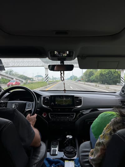 An interior view from a moving vehicle captures a highway scene in the city of Hormiguero, Mexico, under an overcast sky. The image is taken from the rear passenger area, looking forward through the windshield. The driver, identified by forearms and hands gripping a black Honda steering wheel, wears a dark short-sleeved shirt. To the right, a front passenger is visible from the back of their head and shoulders, wearing a green garment and a patterned top.

The dashboard is dark and features a central infotainment screen displaying a navigation map with "1:19" visible. Below the screen are climate controls and other buttons, including "SYNC," "AUTO," and "REAR CONTL." To the right of the steering column is a red push-button start/stop ignition. A gear selector panel with buttons for "P," "R," "N," "D/S," and a switch for "L" and "SNOW" mode is present on the center console. Two cup holders in the console contain a black metal can and a black travel mug. A blue cloth and a yellow object are also visible on the console. A rosary with a crucifix hangs from the rearview mirror, which reflects a masked individual.

Outside the vehicle, a multi-lane highway stretches into the distance. Yellow double lines separate opposing traffic, and white dashed lines mark lanes. On the left, an elevated road structure carries a red compact car traveling in the opposite direction. On the right, further down the highway, a white vehicle proceeds in the same direction. The road is bordered by green trees and bushes. Overhead concrete structures span the highway, and concrete pillars with black and white diagonal hazard stripes are visible along the right shoulder.