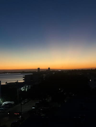 A twilight landscape displays a gradient sky, transitioning from deep blue to soft orange. Below the horizon line, dark silhouettes of buildings are visible, interspersed with faint lights. A body of water reflects the fading sky. Trees line a dark street populated with parked cars and illuminated by sparse streetlights and headlights. The scene suggests a city at dusk viewed from an elevated position.