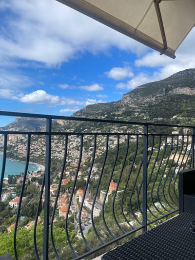 The image shows a view from a balcony in Roquebrune-Cap-Martin, France. The balcony has a black metal railing with a white awning overhead. The railing is curved in a wave-like pattern. The view is of a hillside covered with buildings, trees, and foliage. There is a winding road in the foreground, and a body of water is visible in the background. The sky is blue with scattered clouds. The balcony is empty, with the exception of the corner of a wicker chair. The image was taken during the day.