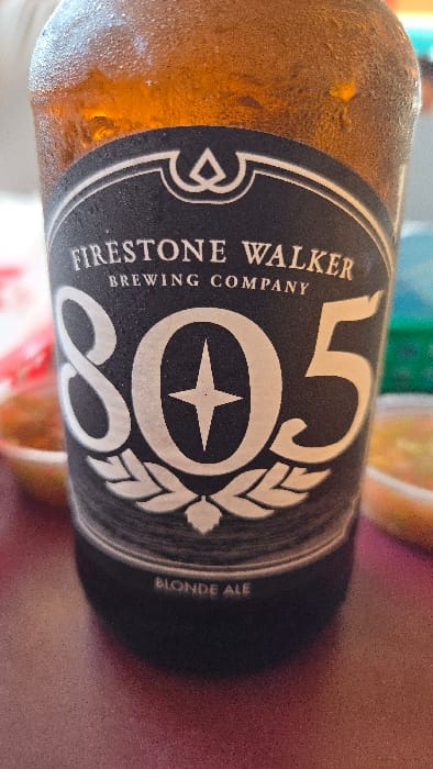 A close-up shot shows a bottle of Firestone Walker Brewing Company's 805 Blonde Ale. The label is prominent, featuring a black background with white lettering. The brand name "Firestone Walker Brewing Company" is clearly visible, as is the large "805" logo. The "Blonde Ale" designation is also present. The bottle has condensation, indicating it is cold. The background is blurred, suggesting shallow depth of field.