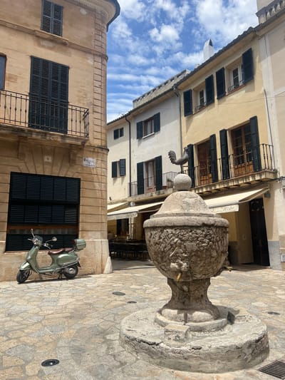 In Pollença, Spain, an outdoor square features a stone fountain with a rooster weather vane. A light green scooter is parked to the left of the fountain. Buildings with closed shutters flank the square, and a street sign for "PLACA DE L'ALMOINA" is visible on the corner of one building. The ground is paved with irregular stone tiles. A few tables and chairs are set up at an outdoor cafe in the background, and a person is seated at one of the tables. The sky is partly cloudy.