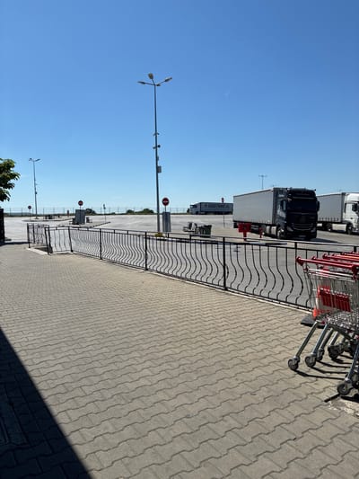 The image shows a sunny outdoor scene, possibly in a parking or loading area, located near Lesovo, Bulgaria. The ground is paved with interlocking concrete blocks. A black, decorative, wave-patterned metal fence separates the paved area from a loading zone containing several large trucks, including a black Mercedes-Benz semi-trailer. To the right of the fence, a row of shopping carts is visible. Light poles with three directional lamps extend upwards towards a clear, blue sky. A thin contrail cuts through the sky in the distance. In the background beyond the trucks, other parking or storage areas are visible, delineated by a wire fence. Stop signs on metal poles are present.