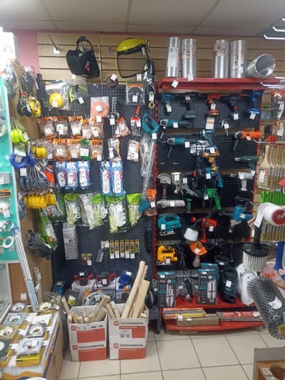 The image depicts a retail store display filled with various tools and hardware items. The scene is likely in Furmanov, Russia, given the presence of Russian signage. On the upper shelves, there are rolls of flexible metal ducting, yellow and black safety helmets, and a black tool bag. Below these, a pegboard wall displays an assortment of tools, including drills, angle grinders, sanders, and spray guns, in various colors like teal, orange, grey, and red.
On lower shelves and to the left, there are rows of packaged electrical extension cords with multiple outlets, coiled ropes in yellow and blue, and various smaller hardware items like wire connectors and clamps. In the foreground, to the right, are cardboard boxes filled with wooden dowels or rods, and on the far right, a large brush attachment for a tool is prominently displayed. The store appears to be well-stocked, with a focus on power tools and construction supplies. The overall environment suggests a typical hardware store or DIY center.