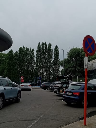 The image depicts a parking lot in Melsele, Belgium, on an overcast day. Several cars are parked, including a light blue SUV in the foreground left, a silver convertible in the mid-ground, and a dark blue hatchback with its trunk open in the right mid-ground. Tall, slender trees line the background. A red sign with a blue circle and a red diagonal cross, indicating no parking, is prominently displayed on the right. Another red and white circular sign with a diagonal red bar and a 'P' symbol is visible on the left. The ground is paved asphalt with visible white parking lines. Charging stations for electric vehicles are visible in the background. There are no people visible in the scene.
