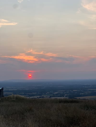 The image shows a sunset over the landscape of Mid Sussex, United Kingdom. The sun, a bright red orb, is low on the horizon, partially obscured by wispy orange and pink clouds. The sky transitions from a pale yellow-orange near the sun to a soft pink and then a light blue at the top of the frame. In the foreground, dry, golden-brown grass covers a hill. To the left, a dark, rectangular structure is partially visible. Below the horizon, a hazy view of rolling hills and fields extends into the distance, with some trees and scattered buildings discernible. The overall scene depicts a serene natural landscape at dusk.