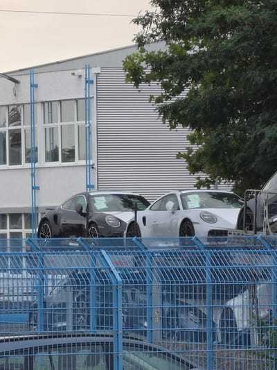 The image depicts a storage area for vehicles, likely a car dealership or a transport hub, located in Ginsheim-Gustavsburg, Germany. The foreground is dominated by a bright blue metal fence, behind which are several vehicles. Two Porsche sports cars are clearly visible on a raised platform. One is black and the other is silver, both appearing to be new and possibly awaiting delivery or sale, as evidenced by stickers on their windshields. In the background, a modern building with a white textured facade and large windows is visible, alongside a section of a building with corrugated metal siding. A large tree with dense green foliage partially obscures the buildings and the sky, which appears overcast. The overall impression is one of an organized, functional space for the storage and display of automobiles within an industrial or commercial setting in Ginsheim-Gustavsburg. There are no people visible in the frame.