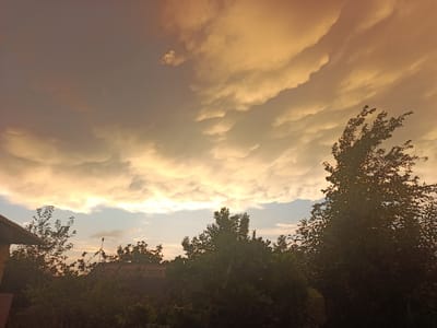 The image depicts a cloudy sky at dusk, with the sun setting behind the clouds. The clouds are illuminated with golden and orange hues, creating a dramatic and picturesque scene. In the foreground, silhouettes of trees and rooftops are visible, suggesting a residential area. The overall atmosphere is calm and serene, with a hint of melancholy associated with the end of the day. The location is Hakkulabad, Uzbekistan, and the time of day is evening. The weather appears to be partly cloudy, with the clouds indicating a potential for rain later in the night. There are no people or text visible in the image.