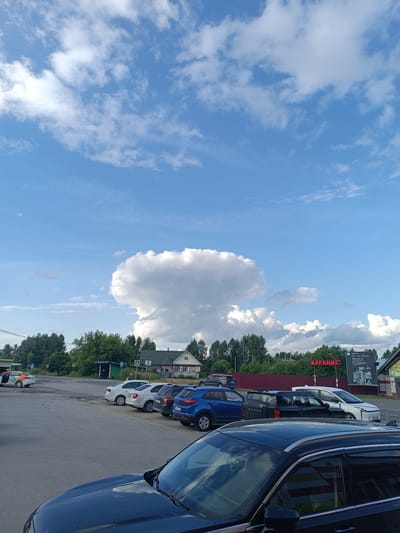 A view of a parking lot in the settlement of Timiryazevo, Russia, is captured under a partly cloudy blue sky. In the foreground, the roof and hood of a dark-colored SUV are visible. Beyond this, several cars are parked in a row on an asphalt surface, including a blue SUV, a black SUV, and a white SUV. Further back, white and grey sedans are parked near a road. Across the road, a few houses with green roofs and trees are situated. A large, mushroom-shaped cloud dominates the sky in the middle distance. On the right side of the image, a red neon sign with the word "БУРЕНИЕ" (Drilling) is displayed above a building. A billboard with text and images is also visible on the right. A person can be seen standing near a bus stop structure in the background.