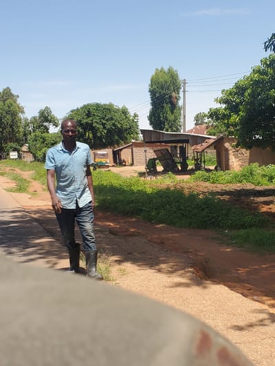 A man stands outside in Mangu, Nigeria. He is wearing a light blue collared shirt and dark pants, with black boots on his feet. The man is facing the camera with his left hand on his hip and his right arm at his side. He appears to be smiling.
The scene is outdoors with a clear blue sky. To the left of the man is a building with a corrugated metal roof, possibly a workshop or storage area, with some machinery visible underneath. Green trees and vegetation surround the building and a dirt path runs alongside the man. In the background, there are more trees and buildings. The time of day appears to be sunny, suggesting daytime. There is a sign on the left side of the image that is partially visible and illegible. The overall setting appears to be rural or semi-rural.