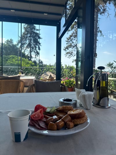 The image depicts a close-up of a table setting at a restaurant in Ognyanovo, Bulgaria, with a focus on breakfast items. The table is covered with a white tablecloth. In the foreground, to the left, is a white paper cup with a dark blue logo and text, possibly indicating a coffee brand. To the right of the cup, a white plate holds a breakfast assortment, including sliced sausages, salami, chopped tomatoes, and cucumber slices. Next to the plate, there is a metal ashtray. Further to the right, a chrome condiment set holds salt and pepper shakers and a bottle of olive oil. In the background, through large glass windows, a scenic view of lush greenery and trees under a bright blue sky is visible. There are also glimpses of outdoor seating areas with wicker chairs and tables. The overall scene suggests a meal taken on a terrace or dining area with an expansive view.