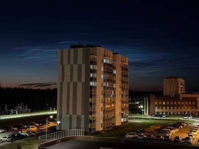 This media file depicts an outdoor scene at dusk or night in Tsiolkovsky, Russia. The primary subjects are residential buildings, a parking lot filled with cars, and a playground. The sky is dark blue, with faint, wispy clouds that appear illuminated, suggesting the presence of noctilucent clouds, a phenomenon often visible at twilight. Two bright points of light, likely stars or planets, are visible in the upper sky, along with a faint streak that could be an airplane.

The architecture of the buildings suggests a modern residential complex. One tall building in the foreground is characterized by a grid-like pattern of alternating gray and cream-colored vertical stripes and numerous windows, many of which are lit from within. In the middle ground, a row of cars is parked on an asphalt surface. The cars vary in color and model, indicating a typical residential parking area. Beyond the parking lot and playground, a dense line of trees forms a dark horizon against the lighter sky. Further in the distance, other buildings are visible, some with illuminated windows.

A playground structure with slides and climbing elements is positioned near the road. A small red goalpost is visible in the foreground, suggesting a sports or recreation area. The overall atmosphere is quiet and still, with the artificial lights of the buildings and streetlights providing illumination against the natural darkness. There is no visible text in the media file. The scene evokes a sense of calm and the transition from day to night in a suburban or semi-urban environment.