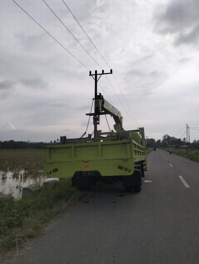 A green truck with a crane attachment is parked on the side of an asphalt road. The truck is facing away from the camera, with its rear visible. The road has white dashed lines. To the left of the road, there is a field with some water, likely a rice paddy, and tall grass. In the background, there are trees and houses. Overhead, power lines stretch across the sky, which is overcast with gray clouds. The scene appears to be in a rural or semi-rural area. The license plate of the truck is partially visible, reading "BH 226 D". The location is Muliengmanyang, Indonesia.