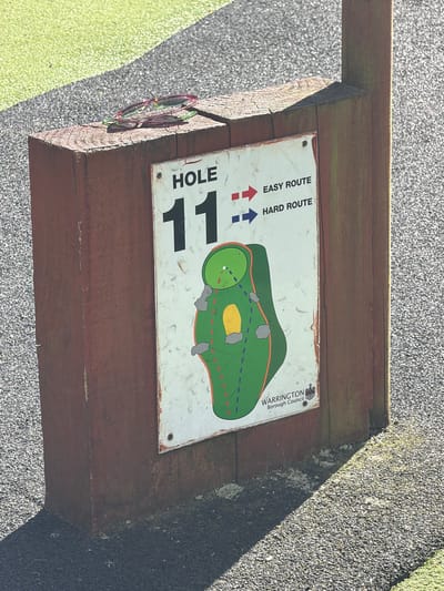 A close-up shot of a wooden stand holding a sign for Hole 11 of a golf course. The sign indicates "Easy Route" and "Hard Route" with colored arrows. A map of the hole is depicted, showing the green, fairway, and hazards. The sign also bears the "Warrington Borough Council" logo, indicating the golf course is located in Warrington, United Kingdom. A pair of sunglasses rests on the side of the wooden stand. The ground around the stand is a mixture of gravel and grass.