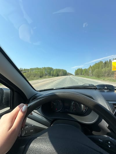 A view from the driver's seat of a car on a road near Керамик, Russia. The sky is blue and clear. The car is moving on an asphalt road, bordered by trees on both sides. A hand is visible on the steering wheel. Sunglasses are on the dashboard. The dashboard instruments include a speedometer showing an unknown speed, and other gauges. A yellow sign is affixed in the upper left corner of the image.