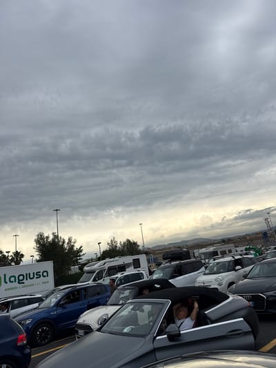 The image shows a parking area in Civitavecchia, Italy, filled with a variety of parked cars, including sedans, SUVs, and campers. A man is seen resting inside a grey convertible car. In the foreground, there is a sign that reads "Iagiusa ARREDI & DESIGN DAL 1960." The background shows a cloudy sky and distant buildings.