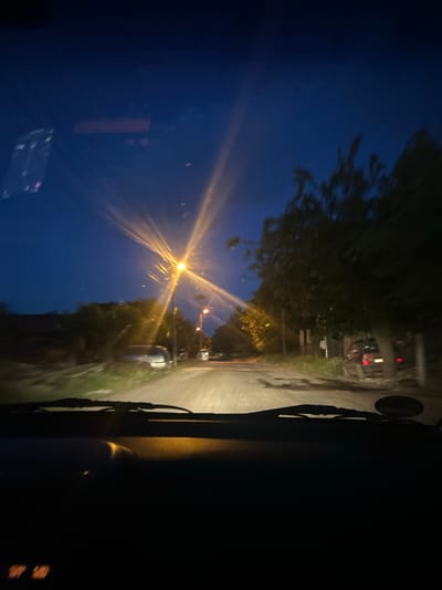 The image shows a view from inside a vehicle, looking out onto a road at night. A street lamp casts a bright glow onto the road and into the sky. Buildings and trees line the road. The road appears wet.
There is not enough information in this image to determine the location in the city of Pishtigovo, Bulgaria.
