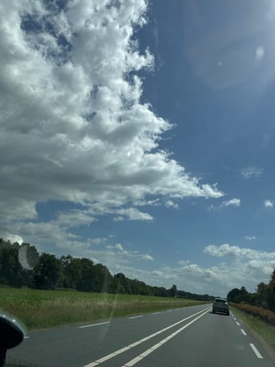 The image depicts a view from a vehicle traveling on a road. The road is asphalt with white lane markings. To the left of the road is a field with green grass and a line of trees. To the right of the road, there is a hedge and more trees. A dark-colored SUV is visible driving in front of the vehicle. The sky is blue with scattered white and gray clouds, and the sun is shining. The scene appears to be in a rural or semi-rural area, possibly Wierden, Netherlands, given the landscape. There are no people visible other than those implied to be in the vehicles.
