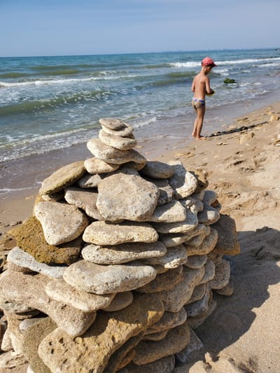 A boy wearing a red cap and patterned swim trunks stands on a sandy beach in Chornomorske, Ukraine, near the edge of the wavy blue sea. He is looking down at something in his hands. In the foreground, a tall, uneven stack of beige and tan stones dominates the view. The sea stretches to the horizon under a clear blue sky, with a distant shoreline visible on the left. The sand is scattered with small pebbles and shells.