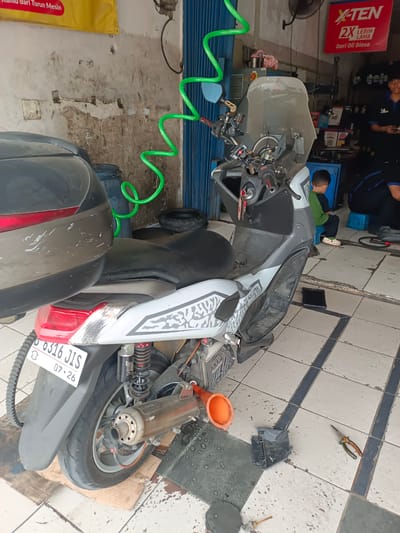 This is a medium shot of a motorcycle repair shop in Serdang Wetan, Indonesia. In the foreground, a white and silver scooter is parked on a cardboard mat. The scooter has a black seat, a windshield, and a silver exhaust pipe. An orange funnel is placed on the ground near the scooter, along with a pair of pliers and some other tools. In the background, two young men are visible. One, wearing a green shirt, sits on a blue stool. The other, in a blue shirt, stands behind a counter, holding a tablet. The shop has a tiled floor and a blue metal shutter door. A green hose hangs from the ceiling. A yellow banner with Indonesian text is displayed above the workspace.