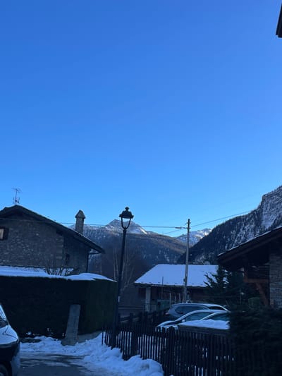 The image captures a serene winter day in a mountain village, presented as if viewed in portrait orientation, although the original file is rotated. A vast, clear blue sky dominates the upper and left portions of the scene. In the background, majestic, tree-covered mountains rise, with their peaks and upper slopes dusted in snow.

In the mid-ground, a prominent, ornate black lamppost stands vertically. To its right, a traditional stone house with a dark roof and a chimney with a pointed top is visible. An antenna is mounted on its roof. Further to the right, behind a dark wooden slatted fence, a silver car is partially visible, parked near buildings with dark roofs, some covered in snow. A dark green hedge, also laden with snow, runs across the mid-ground. The foreground on the lower right shows a small portion of a dark vehicle. Utility lines stretch faintly across the sky against the mountain backdrop.

The ground and all horizontal surfaces are covered in a pristine layer of snow, highlighting the cold, wintry weather. The bright sunlight suggests it is mid-day, casting clear light on the snow and structures. The absence of clouds and presence of snow point to a crisp, clear winter day in a mountainous region like Courmayeur, Italy. No people are visible, and there is no discernible text.