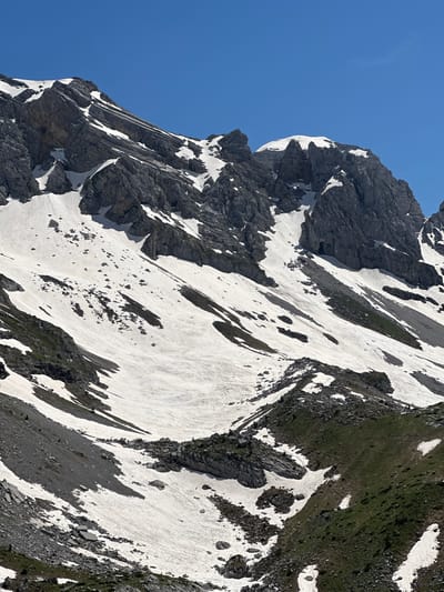 The image displays a panoramic, high-angle view of a rugged, snow-covered mountainous landscape under a clear, bright blue sky in the region of Shalë, Albania. The upper two-thirds of the frame are dominated by steep, grey rocky peaks and ridges, extensively covered with white snow in crevices and on gentler slopes. Layered rock formations are visible on the prominent peak in the upper left, while the peak in the upper right features a rounded, snow-capped summit.

The mid-ground and lower sections reveal vast snowfields interspersed with exposed dark grey rock and scree slopes. Several dark, irregular patches, likely rocks or dirt, are visible within the snow. Towards the lower right, patches of vibrant green vegetation emerge where the snow has melted, indicating the transition to warmer conditions. The scene is devoid of any human subjects, structures, or signs of human activity, presenting a pristine natural environment. The lighting suggests a sunny day, with clear shadows cast by the rocky terrain and snowdrifts.