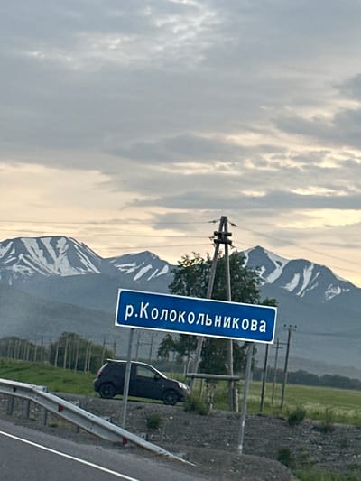 The image depicts a rural scene in the vicinity of Ketkino, Russia. In the foreground, a road with a guardrail is visible, suggesting a transportation route. Beside the road, a dark-colored car is parked. A blue sign with white Cyrillic text, "р.Колокольникова" (which translates to "Kolokolnikova River"), is prominently displayed on a metal pole, indicating a geographical marker. In the background, a row of trees borders a green field, and beyond that, a mountain range with snow-capped peaks under a cloudy sky dominates the horizon. Power lines and utility poles are also present in the scene, suggesting some level of infrastructure. The overall environment appears natural and somewhat remote.