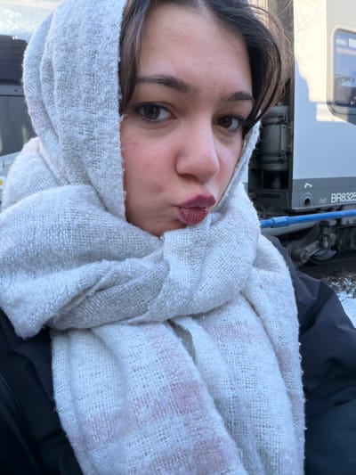 A young adult female is depicted in a close-up selfie taken outdoors in Obermodern-Zutzendorf, France. She has dark hair, dark eyes, and is wearing dark red lipstick, making a puckered lip expression towards the camera. Her head and neck are wrapped in a light-colored, off-white, thick woven scarf featuring subtle pinkish stripe patterns. A dark-colored jacket or coat is visible on her left shoulder. In the background, to her right, a white train carriage or locomotive is partially visible. Markings on the train include "BR8325," a small "ôvô" or "ovo" logo, and a downward-pointing arrow. A blue pipe or hose runs horizontally below the train's main body. The ground in the background appears covered with snow or frost, indicating cold environmental conditions at a railway location.