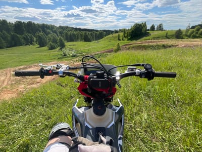 The image depicts a first-person perspective from a stationary dirt bike, specifically a Husqvarna model as indicated by text on the right handlebar grip, overlooking a expansive, verdant valley in Новочувашский, Russia. The foreground features the bike's handlebars, equipped with clutch and brake levers, control modules, a smartphone mounted in a holder displaying "pananode" text, and a red pouch secured below it. A portion of a person's left leg, clad in white attire and a grey and black knee brace, is visible along with a tan-grey gloved hand resting on the bike's fuel tank, which is light-colored with dark grey and white paneling. The ground immediately surrounding the bike is covered in lush green grass interspersed with small white and purple wildflowers.

The midground and background reveal a rolling landscape under a bright blue sky with scattered white cumulus clouds. The valley is characterized by multiple green hills, dense groves of trees (some appearing wind-swept), and clearings of green fields. Visible dirt tracks meander through the terrain, suggesting off-road use, with one prominent track ascending a hill to the right. A small stream or water body is discernible in the lower-left section of the valley. The overall scene indicates a sunny day with clear visibility.