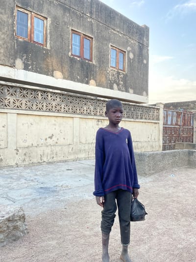 The image depicts a young male child standing in a dusty outdoor environment in Daurawa, Nigeria. The child, with dark skin and short hair, is positioned centrally, facing slightly to the viewer's right with a neutral expression. He wears an oversized, long-sleeved navy blue V-neck sweater with thin red trim and dark grey trousers. His lower legs and feet are bare and coated with light-colored dust or dried mud. He holds a black plastic bag in his left hand, hanging at his side.
In the immediate background is a two-story building with weathered, dark grey concrete or stucco walls showing peeling and discoloration. The upper floor features three equally spaced, rectangular windows with reddish-brown wooden frames and light-colored panes or coverings. Below these windows, a horizontal band of decorative concrete displays a repeating open geometric pattern. To the right of the main building and further back, a rusty, corrugated metal gate or door is partially visible. The ground consists of light brown, compacted dirt and gravel, with scattered lighter-colored concrete or stone fragments. The sky above is a light blue with scattered white clouds.