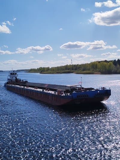 A barge navigates a river under a partly cloudy sky. The vessel is of red and blue color, and appears to be carrying a dark, flat load. The body of water reflects sunlight, creating shimmering effects on the surface. A tree-lined shoreline is visible in the background, with a tall tower extending from the treeline.