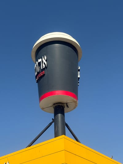 The media file features a close-up, low-angle shot of a cylindrical object against a clear blue sky. The object appears to be a sign or part of a structure, with a dark grey cylinder featuring white and red lettering. The white lettering spells out "SLIC," while the red lettering, partially obscured, seems to read "THE." The cylinder has a beige rim on the left and a red band around its circumference. A black rod extends from the right side of the cylinder, connecting to a bright yellow, angled structure with black lines. This yellow structure is part of a larger object, possibly a building or another sign, given its sharp, geometric form.

The overall impression is that of an outdoor scene under bright daylight, suggested by the vivid blue of the sky and the strong lighting on the objects. The upward-facing perspective emphasizes the scale of the structures against the sky. There are no people visible in the frame. The image was taken in the Bnei Shimon Regional Council, Israel, which is a region known for its varied landscape, including natural reserves and agricultural areas. The context suggests this could be a commercial or public sign within that region. The time of day is likely midday due to the clear, bright sky and lack of shadows that would indicate early morning or late afternoon. The weather appears to be clear and sunny.